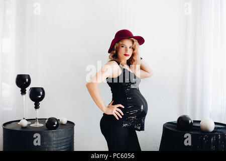 Seitenansicht des trendigen lady in red hat und Schwarz top mit spakling Elemente warten baby posiert zwischen zwei Tabellen mit modernen Möbeln. Ausschnitt auf whit Stockfoto
