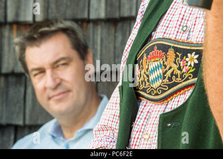 Hausham, Deutschland. 01 Aug, 2018. Markus Soeder (L) der Christlich Sozialen Union (CSU), Ministerpräsident von Bayern, sitzt in der Kreuzbergalm Biergarten hinter dem bayerischen Wappen, die auf das Kostüm eines Teilnehmers in der alpinen Wanderung abgenutzt ist. Wegen der anhaltenden Hitze, in diesem Jahr weniger Menschen nahmen an der jährlichen wichtigsten Alm Inspektion der Alm Management Association. Credit: Lino Mirgeler/dpa/Alamy leben Nachrichten Stockfoto