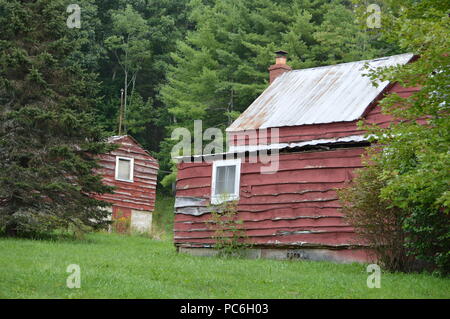 Rustikale rote Haus mit alten Blechdach auf grünem Gras bewachsenen Hügel von immergrünen Bäumen umgeben Stockfoto