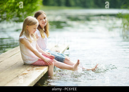 Zwei süße kleine Mädchen sitzen auf einer hölzernen Plattform durch den Fluss oder See Ihre Füße eintauchen in das Wasser an warmen Sommertagen. Aktivitäten für die ganze Familie in Summe Stockfoto