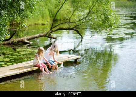 Zwei süße kleine Mädchen sitzen auf einer hölzernen Plattform durch den Fluss oder See Ihre Füße eintauchen in das Wasser an warmen Sommertagen. Aktivitäten für die ganze Familie in Summe Stockfoto