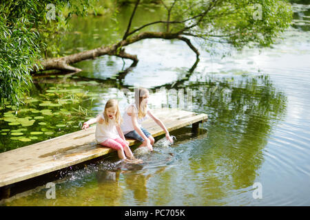 Zwei süße kleine Mädchen sitzen auf einer hölzernen Plattform durch den Fluss oder See Ihre Füße eintauchen in das Wasser an warmen Sommertagen. Aktivitäten für die ganze Familie in Summe Stockfoto