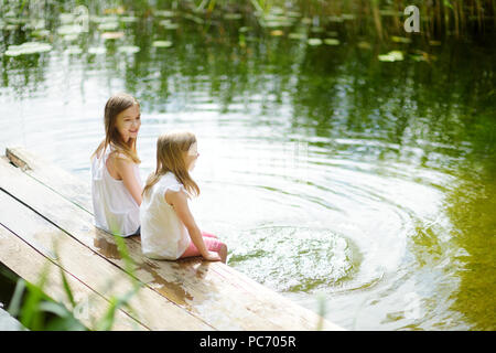 Zwei süße kleine Mädchen sitzen auf einer hölzernen Plattform durch den Fluss oder See Ihre Füße eintauchen in das Wasser an warmen Sommertagen. Aktivitäten für die ganze Familie in Summe Stockfoto