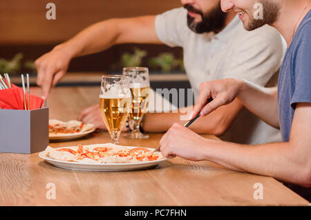 Nahaufnahme der Männer schneiden appetitliche Pizza mit Hilfe von Messer und Gabel. Zwei männliche Freunde haben auch leckeres Bier auf hölzernen Tisch. Konzept der Pizzeria Gastronomie und Küche. Stockfoto