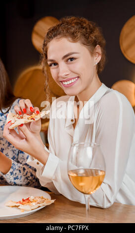 Schöne und bezaubernde junge Frau im Restaurant sitzen und essen lecker Pizza. Ziemlich lockig Frau an der Kamera schauen und lächeln. Glas Weißwein auf dem Tisch platziert. Stockfoto