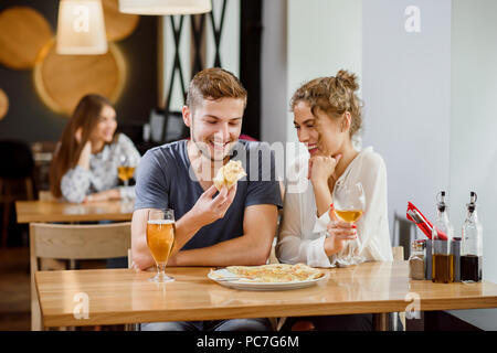 Innenraum der modernen Pizzeria. Süßes Paar sitzt in der Nähe von Tisch, lächeln und posieren. Schöne Frau mit lockigem Haar und schöner Mann essen Pizza. Pizza und Gläser Wein und Bier auf den Tisch. Stockfoto