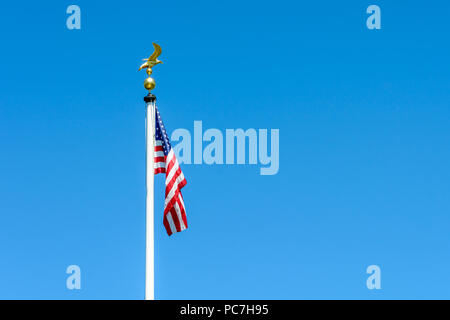 Die Flagge der Vereinigten Staaten von Amerika baumelt am Vollmast auf einem weißen Pol mit einem goldenen Adler auf Ball gegen blauen Himmel gekrönt. Stockfoto