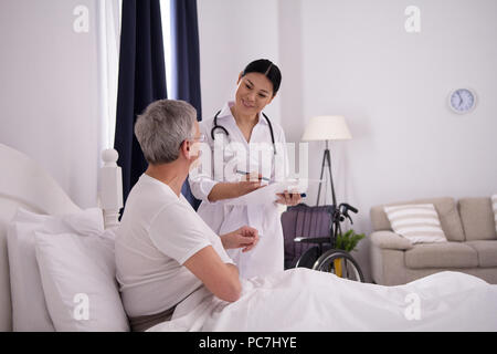 Schöne Krankenschwester, Quick Check auf Patienten. Gut gelaunt freundlich asiatischen Arzt holding Zwischenablage zu prüfen, wie ihre ältere Patienten tut. Stockfoto