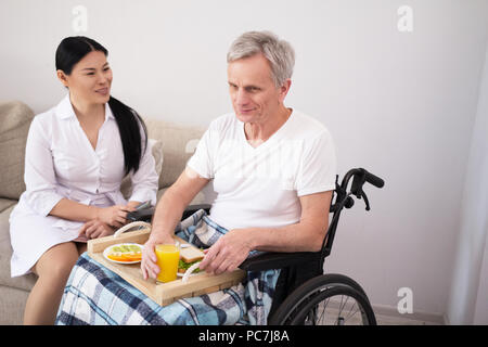 Krankenschwester sitzt neben Mann, während er isst. Schöne asiatische Dame Wiedergabe nach alter Mann, der das Mittagessen auf Essen Fach während der Sitzung in wheelchai Stockfoto