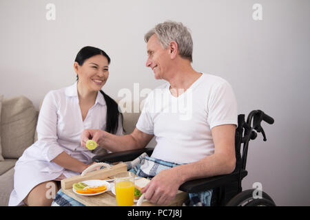 Schönen lächelnden Dame suchen nach Jahren disbaled Mann. Hübsche Weibliche asiatische Krankenschwester lachend, während im Gespräch mit ihrem behinderten älteren Patienten im Rollstuhl. Stockfoto