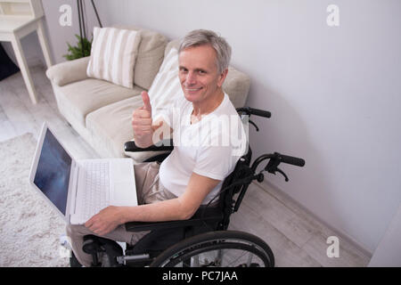 Alte behinderte Menschen das Arbeiten mit Laptop. Alter Mann mit grauem Haar, die Daumen bis zu Kamera beim Sitzen im Rollstuhl mit Computer. Stockfoto