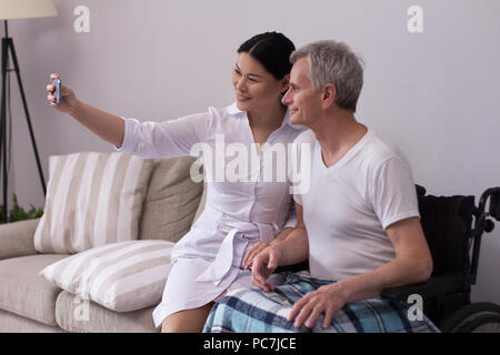 Freundliche Hündin Krankenschwester unter selfie mit Patienten. Schöne asiatische Arzt unter Bild von sich selbst mit alten behinderten Menschen im Rollstuhl per Smartphone. Stockfoto