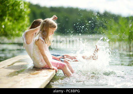 Zwei süße kleine Mädchen sitzen auf einer hölzernen Plattform durch den Fluss oder See Ihre Füße eintauchen in das Wasser an warmen Sommertagen. Aktivitäten für die ganze Familie in Summe Stockfoto