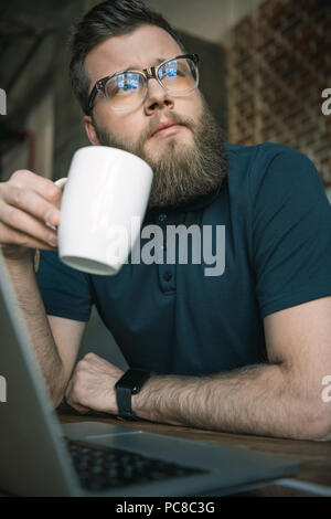 Junge bärtige Mann in Gläser Kaffee trinken während Sie am Tisch sitzen Stockfoto