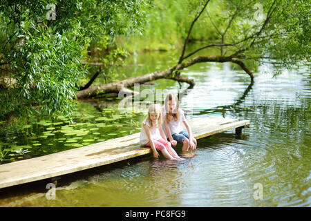 Zwei süße kleine Mädchen sitzen auf einer hölzernen Plattform durch den Fluss oder See Ihre Füße eintauchen in das Wasser an warmen Sommertagen. Aktivitäten für die ganze Familie in Summe Stockfoto