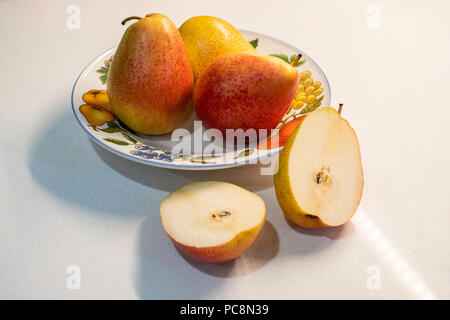 Ganze drei Bartlett Birnen, Pyrus Communis, in einer dekorativen Platte, eine Birne in zwei Hälften geschnitten. Eine weißen Hintergrund Arbeitsplatte. Stockfoto