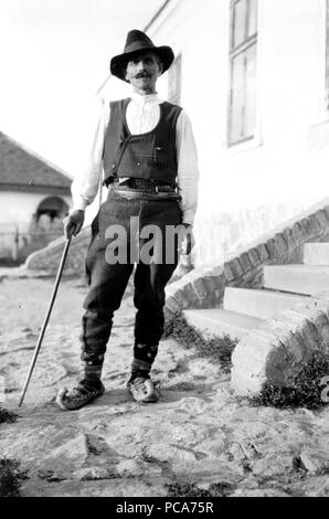 Serbische Mann mit Hut, Hemd, Hose, Gürtel und spitzen Schuhe mit Zuckerrohr stehen außerhalb der Gebäude in Jugoslawien c 1918-1924 Stockfoto