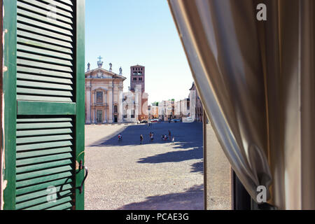 Der Blick aus dem Fenster mit grünen Fensterläden aus Holz und schönen Gardinen von der Sonne auf dem Platz mit Kindern beim Spielen in der italienischen Stadt Ma Stockfoto