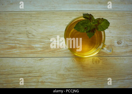 Ein Glas Becher mit Griff mit goldenen Tee mit Minze Blätter und Blumen steht auf einer Untertasse und auf einem Holztisch im Sommer Stockfoto