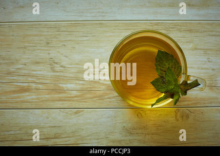 Ein Glas Becher mit Griff mit goldenen Tee mit Minze Blätter und Blumen steht auf einer Untertasse und auf einem Holztisch im Sommer Stockfoto