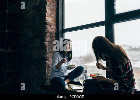 Zwei freundliche junge Frauen haben Abendessen sitzen auf der Fensterbank zu Hause Stockfoto