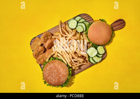 Holzbrett mit unterschiedlichen Fast food: Burger, Chicken Nuggets, Pommes frites auf gelben Hintergrund. Ansicht von oben mit Platz kopieren. Muster. Flach. Stockfoto