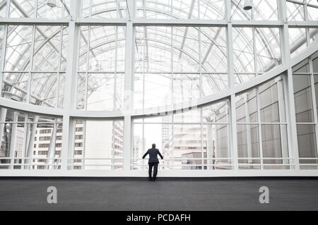 Ein Mann stand in einem offenen Raum in einem gläsernen Atrium in einem Bürogebäude, stützte sich auf ein Geländer, Ansicht von hinten. Stockfoto