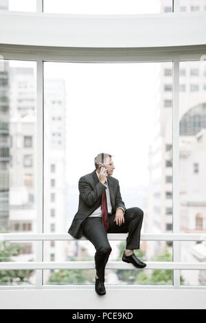Ein kaukasischer Geschäftsmann mit seinem Handy, während er auf dem Geländer an der Basis von einem großen Fenster in einer Convention Center. Stockfoto