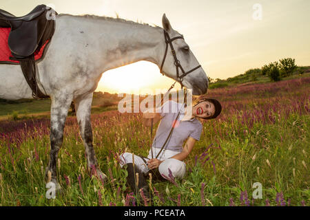 Schöne lächelnde Mädchen Jockey stand neben ihr weißes Pferd Tragen besonderer Uniform auf einen Himmel und grünen Feld Hintergrund auf einen Sonnenuntergang. Stockfoto