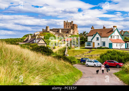 Zwei Frauen gehen Hund von Bamburgh Strand Straße nach Bamburgh Dorf, mit Bamburgh Castle im Hintergrund. Bamburgh, Northumberland, Großbritannien. Stockfoto