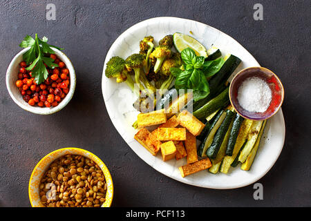 Gesunde vegetarische Nahrung. Gebackene Gemüse (Zucchini und Broccoli), gebratener Tofu, Käse, Braten, Kichererbsen und grüne Linsen auf dunklem Hintergrund, Ansicht von oben Stockfoto