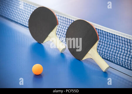 Orange Kugel für Tischtennis und zwei hölzerne Schläger von roten und schwarzen Farbe auf einem blauen Tabelle mit einem Raster Stockfoto