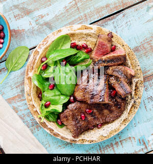 Close up Rindersteak vom Grill mit Spinat Blätter und Granatapfel in der Platte auf Blau Holz- Tabelle, Ansicht von oben Stockfoto