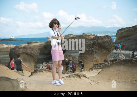 Chinesische Mädchen mit Sonnenbrille und Bob Haircut, tragen weiße Turnschuhe, ein selfie mit einem selfie Stick in ein geopark, Mit dem Meer im Rücken. Stockfoto