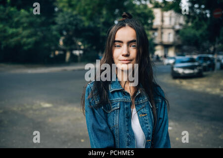 Jugendlich Mädchen in Blue Jeans Jacke mit Kamera gekleidet und lächelnd gegen das Grün der Bäume und auf der Straße. Portrait von glücklich schöne junge Frau in der Stockfoto