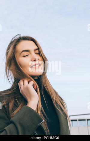 Junge Frau mit stilvollen khaki Fell und schwarzen Rollkragenpullover genießen Herbst Tag. Portrait von Mädchen mit langen braunen Haaren lächelnd und schließen die Augen outdoo Stockfoto