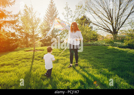 Schöne und stilvolle rothaarige Mama in eine weiße Bluse spielen mit ihren kleinen süßen Sohn in einem Sommer sonnigen Park mit einem Kite Stockfoto