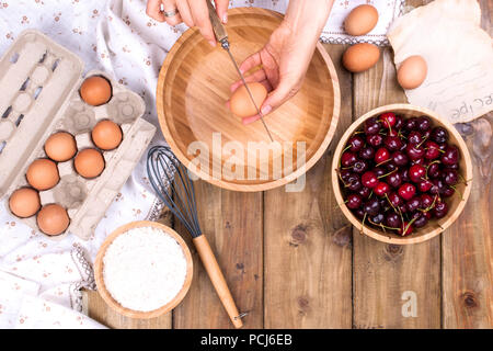 Weibliche Hände brechen das Ei in eine Schüssel geben. Zubereitung von Teig für eine Torte mit einer Kirsche. Sommer hausgemachtes Gebäck. Ansicht von oben. Platz kopieren Stockfoto