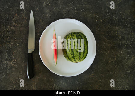 Eine halbe Wassermelone auf einem weissen Teller, neben einem Messer, beide Kompositionen machen jeweils zehn Stockfoto