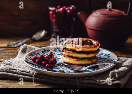 Russische Quark Pfannkuchen mit Schokolade Topping und gefrorene Kirschen. Teekanne mit Löffeln und Glas von Beeren für den Hintergrund. Stockfoto