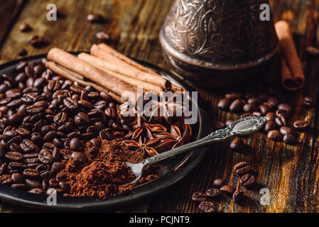 Kaffee Bohnen mit Spooonful gemahlenen Kaffee, Zimtstangen und Chinesischer Sternanis auf der Metallplatte. Bohnen verstreut auf Holztisch und Cezve auf der Rückseite Stockfoto