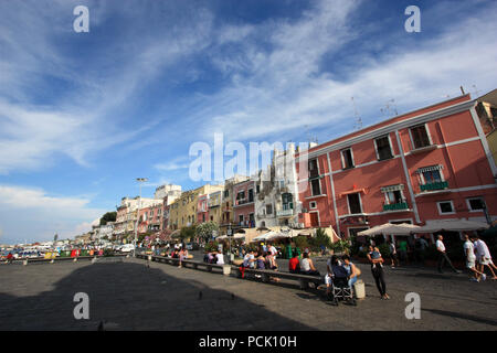 Viele Touristen entlang der Promenade auf Procida, Italien bummeln, mit bunten Häusern in verschiedenen Pastelltönen im Hintergrund gemalt Stockfoto