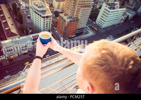 In der Nähe der Hände des Menschen Holding entfernt Pappbecher mit Morgen heiße Getränke - Kaffee oder Tee mit inspirierenden Blick auf abstrakte Stadt Hintergrund bei Sonnenaufgang oder Sonnenuntergang. Ansicht von oben. Weiche selektiven Fokus. Platz kopieren Stockfoto