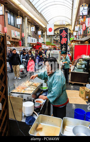 : Kuromon Ichiba, Küche essen Osaka's Markt. Blick entlang Arcade mit Stall im Vordergrund verkauf Oden, typische Winter essen. Markt überfüllt mit Menschen. Stockfoto