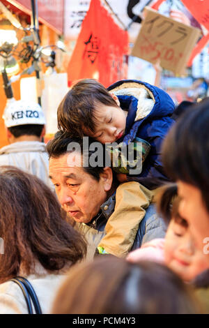 Japanische junge Kind, Junge, schlafend auf seinem Vater zurück, so wie er ist durch einen belebten Markt in der Tageszeit durchgeführt. Candid Schuß an: Kuromon Ichiba, Osaka. Stockfoto