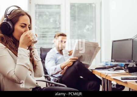 Mit Kopfhörern am Schreibtisch sitzen während der Pause und mit Handy während man liest Zeitung Geschäftsfrau Stockfoto