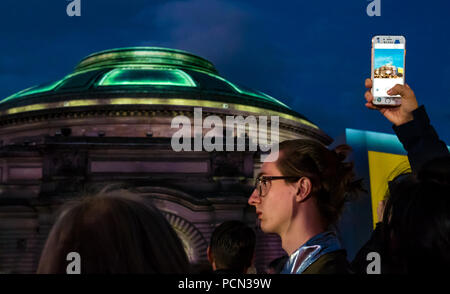 Edinburgh, Großbritannien. 3. Aug 2018. Aberdeen Standard Investitionen Sponsoren das Edinburgh International Festival 2018 Eröffnung fünf Telegramme mit einer Masse in Festival Square. Feiern Schottlands Jahr der jungen Menschen und auf das hundertjährige Jubiläum des Ende des Großen Krieges reflektieren mit beauftragt, Musik von Anna Meredith und digitale Projektion auf die Usher Hall von 59 Produktionen, die im Freien Fall wird durch Telegramme von jungen Soldaten im Jahr 1918 gesendet. Eine der Sängerinnen, die noch in der zweiten Akt ein Feld Postkarte genannt zu singen, während eine Person ein Foto auf einem Mobiltelefon erfolgt Stockfoto