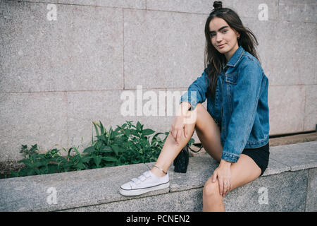 Portrait von glücklichen Mädchen mit langen dunklen Haaren sitzen in der Nähe von grauen Gebäude am Sommer, der Tag. Teen Frauen in Stadt posing Legere Outfit: kurze Hose, Blau je Stockfoto