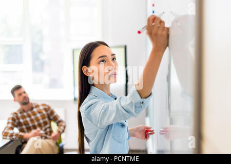 Junge asiatische Schüler Mädchen Schreiben auf dem Whiteboard während der Lektion mit unscharfen Lehrer im Hintergrund Stockfoto