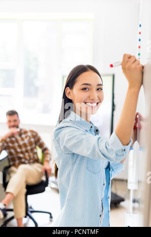 Gerne asiatische Schüler Mädchen Schreiben auf dem Whiteboard während der Lektion mit unscharfen Lehrer im Hintergrund Stockfoto